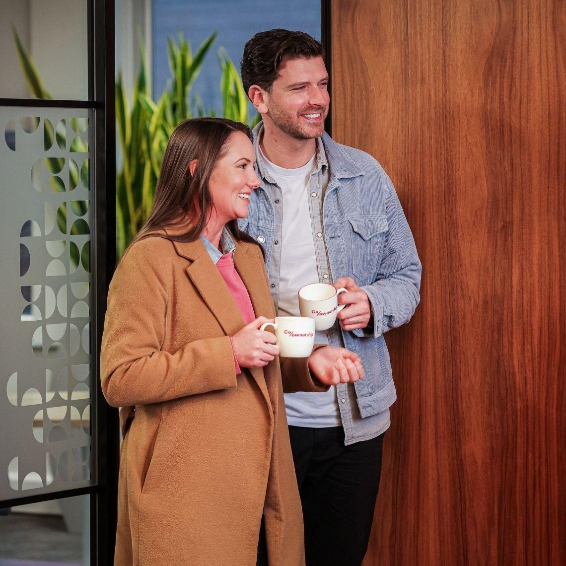 man and woman smiling holding branded mugs standing in doorway of office