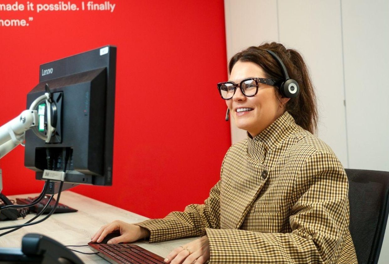 Woman in patterned coat, wearing glasses and a headset, smiling at her computer
