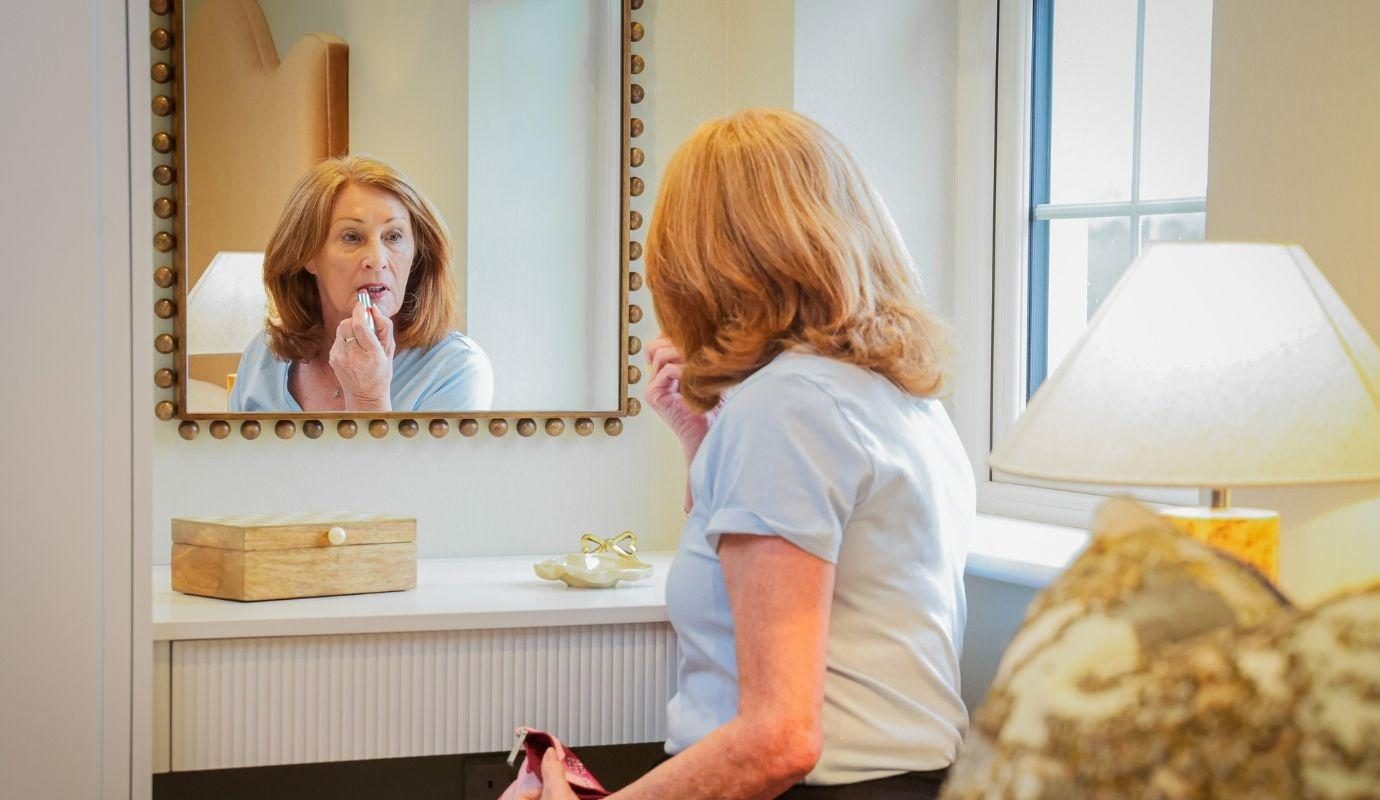 Woman putting on lipstick sitting at bedroom vanity with mirror reflection on camera