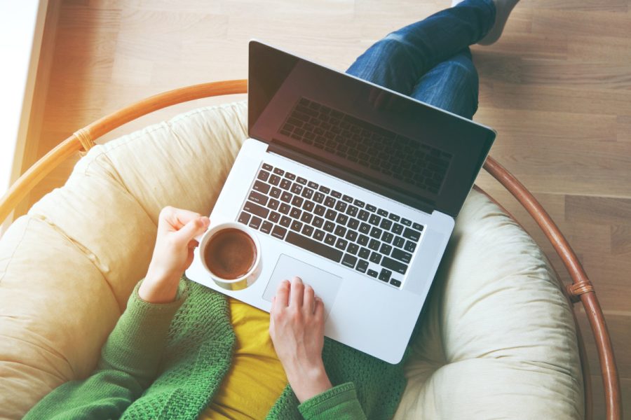 Person sitting in a cushioned chair using a laptop, holding a cup of coffee, viewed from above.
